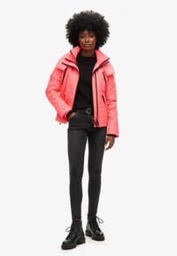 Lys rosa polstret jakke med høj krave, sort trøje underneath, sorte stramme jeans og sorte snørebodder. Glat tekstur, moderne design.