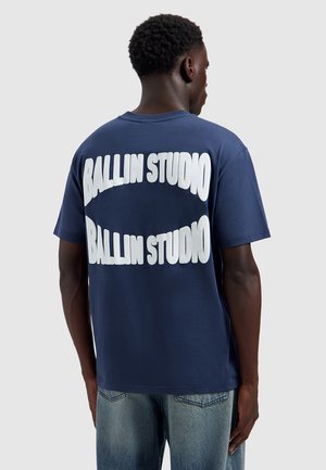 Man die een donkerblauw T-shirt draagt met "BALLIN STUDIO" twee keer in grote witte letters op de achterkant, gecombineerd met lichtblauwe spijkerbroek.