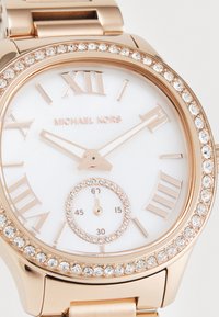 Reloj de metal en tono dorado con un dial blanco, marcadores de hora en números romanos y un bisel adornado con cristales. Presenta un pequeño sub-dial para los segundos.