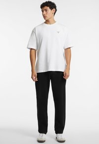 Weißes Baumwoll-T-Shirt mit kurzen Ärmeln und einem kleinen roten Logo auf der linken Brust, kombiniert mit schwarzen Hosen und weißen Turnschuhen.