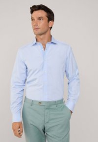 Lichtblauwe blouse met lange mouwen en een button-down kraag, gemaakt van een gladde stof. Gecombineerd met lichtgroene broek, met een knoopdetail.