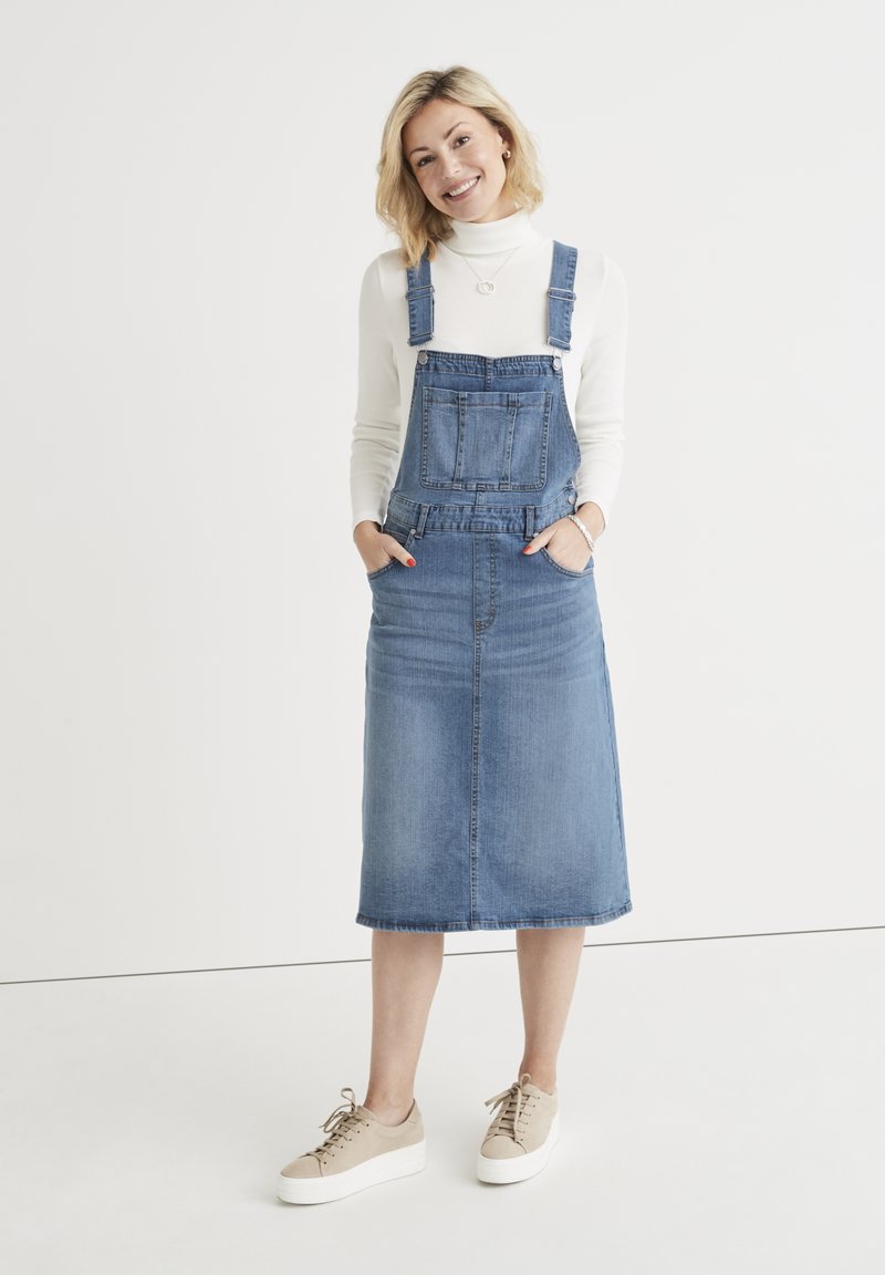 Denim overall dress in light blue with a straight cut, front pocket, adjustable straps, and a knee-length hem. Worn with beige sneakers.