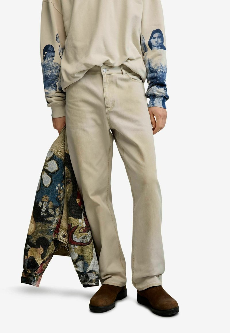 Person wears beige pants and sweatshirt with blue graphic faces on sleeves, holding a multicolored patterned jacket, paired with brown shoes.