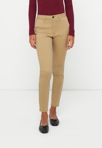 Femme portant un pantalon beige slim-fit, un haut bordeaux à manches longues et des ballerines noires, se tenant devant un fond blanc.