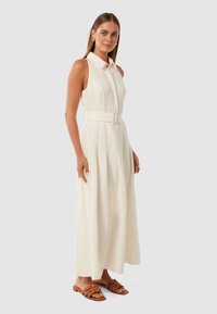 Vestido longo, sem mangas, branco, com gola, cintura cingida por um cinto e saia plissada. Usado com sandálias rasteiras castanhas. Textura de tecido suave.