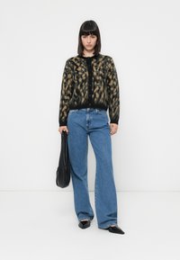 Mulher vestindo um cardigan de estampado de leopardo, jeans azuis de perna larga, sapatos pretos de bico fino, segurando uma bolsa preta, em pé contra um fundo branco simples.
