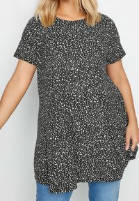 Svart tunika med vitt prickigt mönster, korta ärmar och avslappnad passform; mjukt tyg med subtil textur, kombinerad med blåjeans.