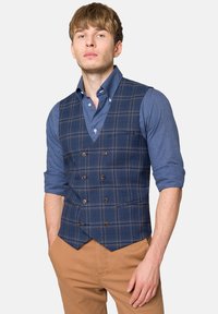 Granatowa kamizelka w kratkę z podwójnym zapięciem, noszona na niebieskiej koszuli zapinanej na guziki. Ma brązowe guziki i jedną kieszeń na piersiach. Beżowe spodnie.