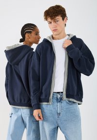 Námořnická mikina na zip s šedými detaily, vyroben�á z měkké tkaniny. Má volný střih a velké klokaní kapsy. Modely nosí džínové kalhoty.