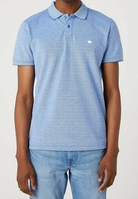 Polo bleu clair en tissu texturé, avec une patte de boutonnage, des manches courtes et un petit logo blanc sur la poitrine.
