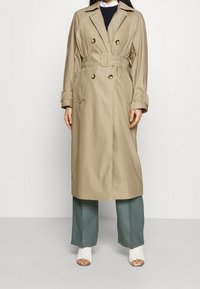 Trench coat beige con vita regolabile, design doppiopetto e grandi bottoni. Presenta tasche laterali e colletto. Abbinato a pantaloni a gamba larga.