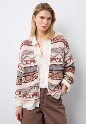 Vrouw met blond haar, gekleed in een gebreid vest met patroon over een witte blouse en een bruine broek, staand met één hand in haar zak tegen een effen achtergrond.