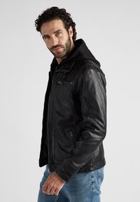 Zwarte leren jas met rits aan de voorkant en twee zijzakken. Bevat een capuchon en gestructureerde mouwen. Model draagt een spijkerbroek.