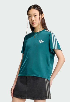 Jonge vrouw met lang gevlochten haar draagt een turquoise Adidas-shirt met witte strepen en een zwarte rok met zijstrepen, staat met haar hand op haar heup.