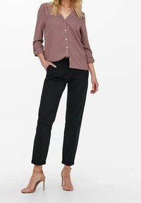 Camicia button-up viola chiaro con maniche lunghe e polsini arrotolati, abbinata a pantaloni neri a sigaretta e sandali con tacco nudo.