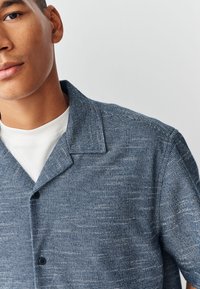 Overhemd van een gestructureerde blauwe stof, met een spreidkraag, knoopsluiting en een ontwerp met korte mouwen, gedragen over een witte ronde hals.