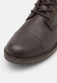 Brown leather shoe with a textured finish, round toe, and stitched accents. Features laces and brass-colored eyelets.
