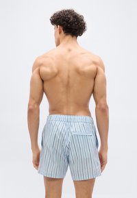 Άνδρας με σγουρά  μαλλιά, χωρίς πουκάμισο, φοράει ανοιχτό μπλε και λευκά κάθετα ριγέ σορτς, στέκεται με την πλάτη προς το μέρος μας σε λευκό φόντο.