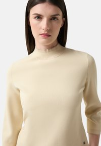 Beige Rippstrickpullover mit hohem Kragen und Dreiviertelärmeln. Verfügt über eine glatte Textur und einen dezenten Logoakzent nahe dem Saum.