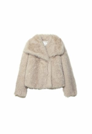 Femme portant une veste en fausse fourrure beige, une chemise rayée, un pantalon marron à jambes larges, des sandales à plateforme, tenant un sac à main marron foncé contre un mur blanc.