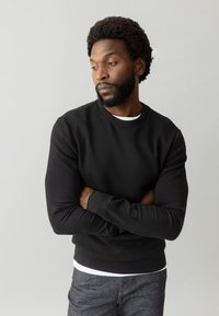 Felpa nera con collo a giro, maniche lunghe e polsini a coste. Abbinata a una maglietta bianca, su uno sfondo neutro.