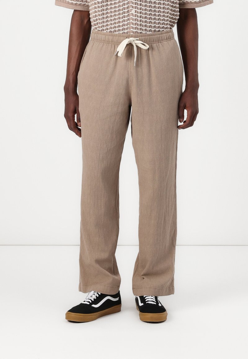 Beige, textured, relaxed-fit pants with an elastic waistband and drawstring. Paired with black sneakers featuring white accents.