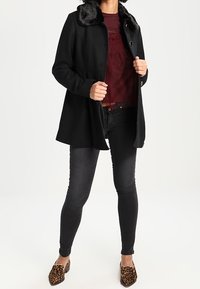 Cappotto nero con colletto in pelliccia sintetica, abbinato a un top bordeaux e jeans neri. Presenta una vestibilità su misura e bottoni sul davanti.