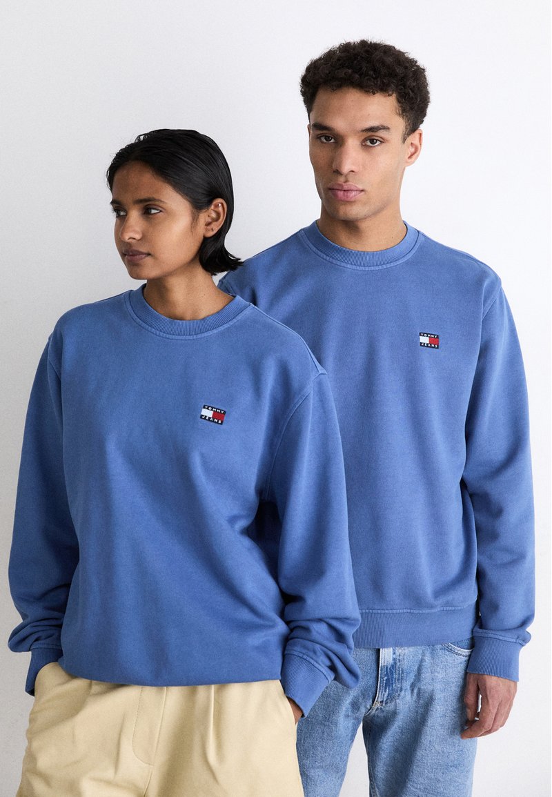 Dos personas llevan sudaderas de cuello redondo a juego en un color azul claro. Cada una tiene un pequeño logo multicolor en el pecho.