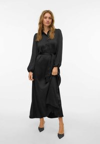 Melna satīna gara kleita ar pogām priekšpusē, gariem pūkainiem piedurknēm, sasietu vidukļa jostu un plūstošu svārku. Komplektā ar punktainiem papēžiem.