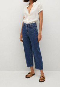 Camisa blanca ligera de mangas cortas, combinada con vaqueros azul de tiro alto y holgados, y sandalias deslizables marrones. Estilo minimalista.
