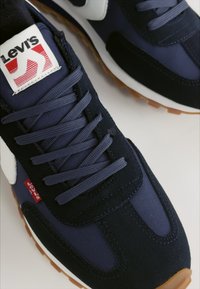 Sötétkék tornacipő velúr és nylon anyagból, lapos fűzőkkel, gumitalppal, és Levi's logóval ellátott címkével a nyelvén.