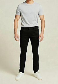 Homme portant un t-shirt gris clair à manches courtes, un pantalon noir ajusté et des baskets blanches, debout devant un fond clair uni.