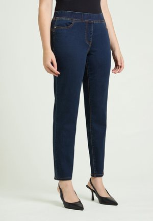 Kvinna som bär mörkblå slim-fit-jeans och svarta spetsiga slingback högklackade skor, stående mot en enfärgad bakgrund.