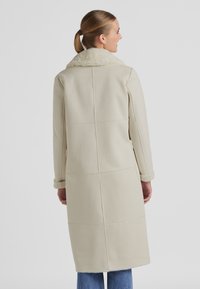 Langer, cremefarbener Mantel mit einem weichen Pelzkragen, gepaneltem Design und umgeschlagenen Ärmeln. Die Rückansicht zeigt eine gerade Silhouette und eine weiche Textur.