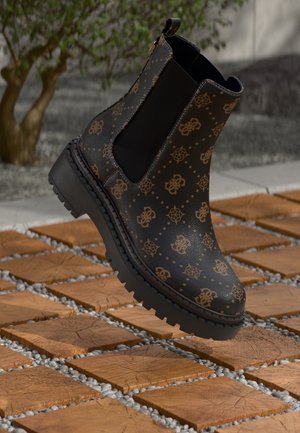 Botte Chelsea noire avec motif logo marron, semelle épaisse, panneaux latéraux élastiques et boucle au talon, présentée sur un pavé en bois et galets.