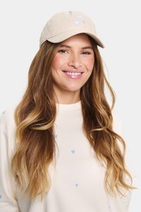 Bézs baseballsapka világoskék szív hímzéssel, világos pulóverrel párosítva, amely apró kék szívmintákat tartalmaz. Hosszú, hullámos haj látható.