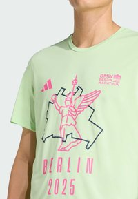 Camiseta atlética verde claro de mangas cortas con elementos gráficos en rosa, que incluyen una figura y el texto "BERLÍN 2025," hecha de material transpirable.