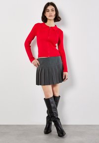 Maglione rosso a coste con zip e colletto, abbinato a una gonna a pieghe grigio scuro a righe. Il modello indossa stivali neri al ginocchio.
