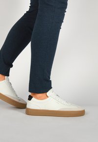 Baskets en cuir blanc avec des semelles en caoutchouc beige, des accents de talon noirs et des lacets blancs ; associées à un pantalon fitted bleu foncé.