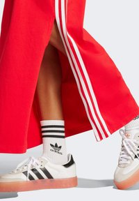 Czerwona spódnica sportowa z białymi bocznymi paskami, w połączeniu z białymi i czarnymi sneakersami z trzema paskami i przezroczystą podeszwą oraz w skarpetkami w paski.