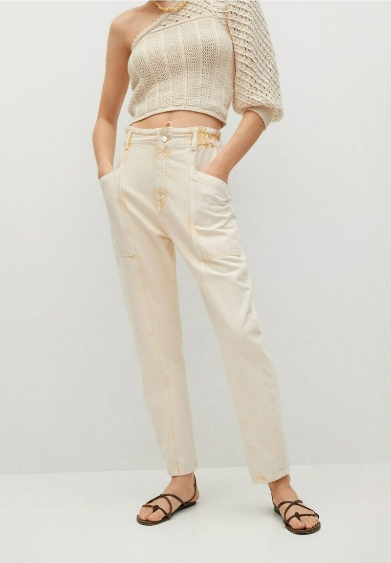 Beige crop-top dengan desain rajutan berbentuk tekstur, dipadukan dengan celana tapered beige muda yang memiliki saku depan dan detail jahitan. Sandal tali cokelat.