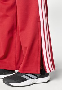 Piros tréningnadrág fehér oldalsávokkal és résszel, fekete sportcipővel kiegészítve, amelyen szürke részletek és texturált felület található.