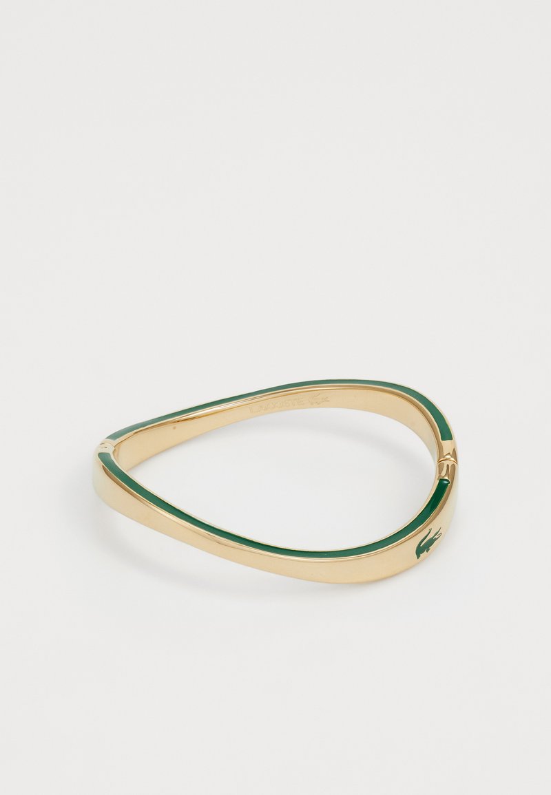 Kuldtono bangles käevõru, millel on roheline emailiga vooder ja Lacoste logo aksent. Sile tekstuur ja kitsenev, nurga kujuline disain.