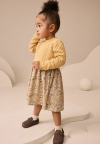Pull en laine jaune avec motif en losanges, robe à imprimé floral avec des couleurs pastel, chaussures brunes à enfiler, et chaussettes blanches.