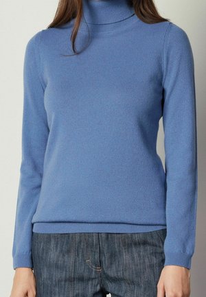 Femme portant un pull à col roulé bleu clair ajusté et un jean bleu foncé sur un fond uni.