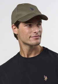 Gorra de béisbol verde oliva con un logo bordado en azul marino. Se lleva con una camiseta negra de manga corta que presenta un pequeño logo colorido en el pecho.