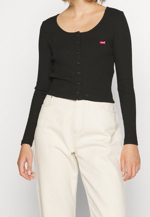 Czarna prążkowana bluzka z długim rękawem zapinana na guziki z czerwonym logo Levi's, w połączeniu z jasnobeżowymi spodniami z wysokim stanem, na osobie na jednolitym tle.