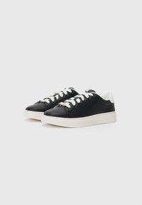 Des sneakers en cuir noir avec une semelle en caoutchouc blanche, des lacets blancs, et un design minimaliste avec un branding sur le côté et des accents contrastants au talon.