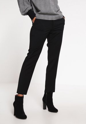 Osoba ubrana w czarne, dopasowane spodnie do kostek oraz czarne, na obcasie botki, z rękami w kieszeniach, ubrana w szarą, kratkowaną bluzkę.