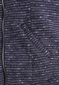 Marineblauwe jas met ritssluiting, gebreid patroon met witte spikkels, zijzak, en geribbelde manchetten en zoom voor een stevige pasvorm.